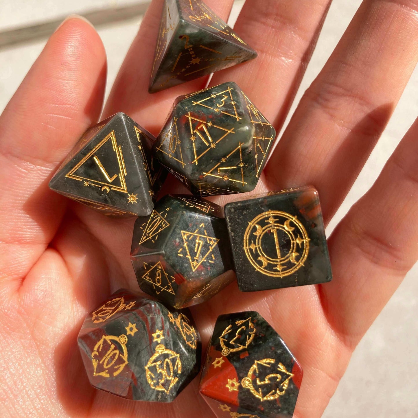 A carefully held Bloodstone Gemstone Engraved Dice Set in natural daylight, highlighting the stone's deep green hues and sharp edges against skin tones