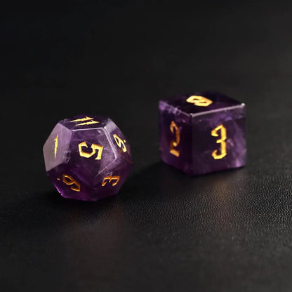Two purple amethyst polyhedral dice showing gold-infill numbers and moon motifs, high-contrast visual on dark leather.